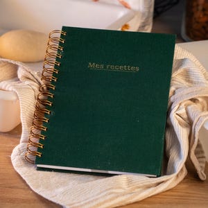 Peut inclure: Un carnet à spirales vert foncé avec une reliure en fil métallique doré. La couverture porte l'inscription "Mes recettes" en lettres dorées. Le carnet est posé sur un tissu texturé de couleur claire sur une surface en bois.