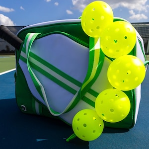Puede incluir: Una bolsa de pickleball verde y blanca con un asa negra y varias pelotas de pickleball amarillas. La bolsa tiene rayas verdes y blancas y una correa verde. Las pelotas de pickleball son de color amarillo brillante con múltiples agujeros. El fondo es una cancha azul con cielo azul.