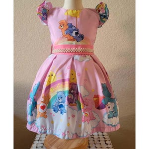May include: A pink dress featuring Care Bears characters, rainbows, and clouds. The dress has a pearl-beaded belt, puff sleeves, and a pleated skirt. The bodice has a Care Bears graphic. The dress is displayed on a mannequin.