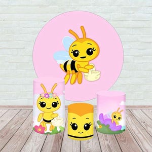 May include: A collection of bee-themed party decorations. The set includes three cylindrical stands and a large pink circle backdrop, all featuring cartoon bee illustrations in yellow, black, and white. The stands vary in size and design.