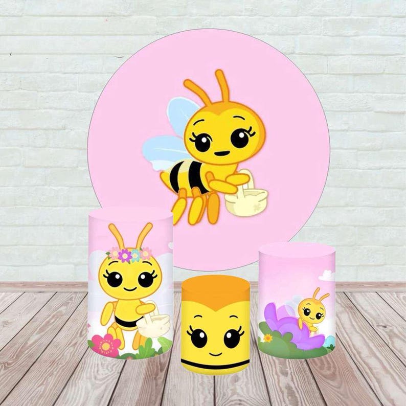 May include: A collection of bee-themed party decorations. The set includes three cylindrical stands and a large pink circle backdrop, all featuring cartoon bee illustrations in yellow, black, and white. The stands vary in size and design.
