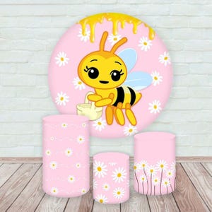May include: A pink backdrop with a cartoon bee and daisy accents, along with three matching pink cylindrical pedestals. The bee is yellow and black, holding a basket. The pedestals are decorated with daisy patterns.