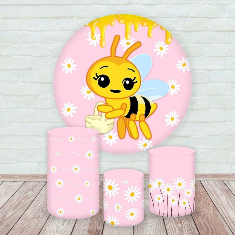May include: A pink backdrop with a cartoon bee and daisy accents, along with three matching pink cylindrical pedestals. The bee is yellow and black, holding a basket. The pedestals are decorated with daisy patterns.