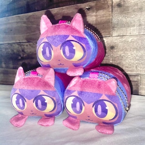 May include: Three plush cat-shaped coin purses in pink, purple, and blue. Each has large, stylized eyes, small ears, and a zipper closure with a silver chain. The purses are stacked on top of each other.