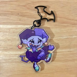 Könnte beinhalten: Ein Acryl-Schlüsselanhänger mit einer Cartoon-Narrenfigur mit hellblauer Haut, einer violetten und dunkelvioletten Narrenkappe und einem dunkelvioletten Outfit. Die Figur hat ein breites Grinsen und ist an einem schwarzen, fledermausförmigen Metallverschluss befestigt.