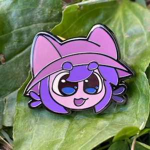 May include: A purple and pink enamel pin featuring a cartoon character with a cat-like hat. The character has large blue eyes, a small smile, and purple hair. The pin has a black outline and is set against a green leaf.