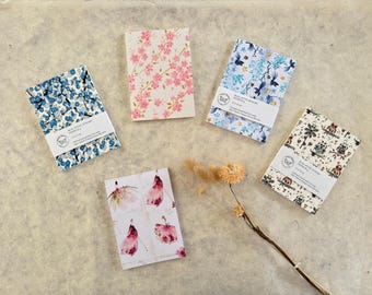 Peut inclure: Cinq petits carnets avec des motifs floraux et à motifs. Les carnets présentent divers motifs, notamment des baies bleues, des fleurs de cerisier roses, des oiseaux bleus et des illustrations fantaisistes. Chaque carnet a une bande blanche avec du texte et un logo papillon.