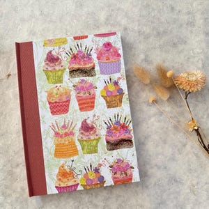Peut inclure: Un cahier à couverture rigide avec un motif de cupcakes. La couverture présente un motif de cupcakes colorés avec diverses garnitures et bougies. La tranche est de couleur brun rougeâtre uni. Le cahier est sur une surface texturée et claire.
