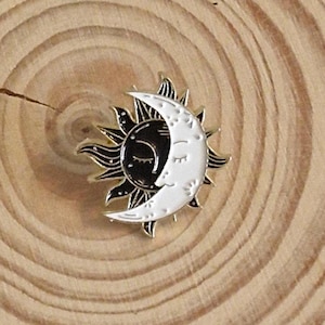 Può includere: Uno spillo in smalto dorato con un sole nero e una luna crescente bianca, con la luna che sovrappone il sole. Il sole ha un viso con gli occhi chiusi e la luna ha un viso con gli occhi chiusi. Lo spillo è su una superficie di legno.