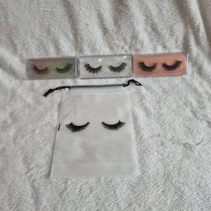 Op de afbeelding: Een assortiment valse wimpers in doorzichtige plastic doosjes, met een witte trekkoordtas met een zwart wimperontwerp. De doosjes zijn gerangschikt op een wit, getextureerd oppervlak. De wimpers variëren in stijl.