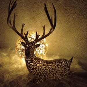 Peut inclure: Une figurine décorative de cerf avec un motif ajouré complexe, placée devant un globe lumineux. Le cerf a de grands bois et repose sur une surface blanche et duveteuse. L'éclairage chaleureux crée une ambiance cosy.