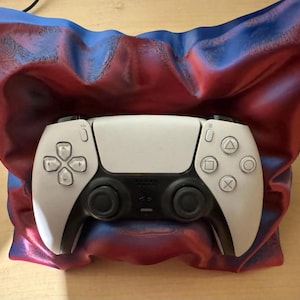 Könnte beinhalten: Ein weiß-schwarzer Videospiel-Controller liegt in einem rot-blau-violetten Halter. Der Controller hat ein symmetrisches Design mit Tasten und Joysticks. Der Halter hat ein drapiertes Stoffaussehen.