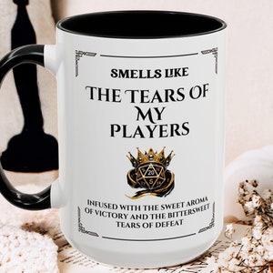 May include: White ceramic mug with a black handle. The mug has a black rim and a design that reads "Smells Like The Tears Of My Players" with a gold crown and dice graphic. The text below the graphic reads "Infused with the sweet aroma of victory and the bittersweet tears of defeat."
