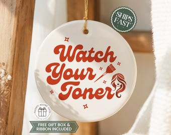 May include: A round white ceramic ornament featuring the red text Watch Your Toner, a red hair dye brush icon, and small red star accents. The ornament hangs from a gold string and includes a circular hole at the top for suspension.
