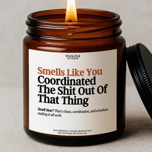 May include: A lit candle in an amber glass jar with a black lid. The white label reads: Wick'd Lit BY VALENTI, Smells Like You Coordinated The Shit Out Of That Thing, Smell that? That's chaos, coordination, and somehow making it all work. 4oz.