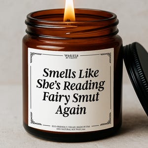 May include: A lit candle in an amber glass jar with a black lid. The white label features a decorative border and black text that reads: Wick'd Lit BY VALENTI, Smells Like She's Reading Fairy Smut Again, ECO-FRIENDLY | VEGAN | MADE IN USA 100% NATURAL SOY WAX | 4oz.