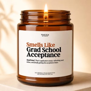 Op de afbeelding: Een brandende kaars in een amberkleurige glazen pot met een wit etiket. Het etiket vermeldt Wick'd Lit by Valenti, Smells Like Grad School Acceptance, met beschrijvende tekst over sollicitatieessays en toelatingsbrieven, plus 113g.