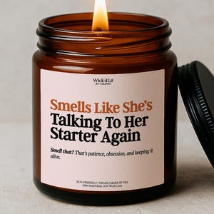 May include: A lit, amber-coloured glass jar candle with a black lid. The candle has a white label with the text "Smells Like She's Talking To Her Starter Again." The candle is made with 100% natural soy wax and is 113 grams.