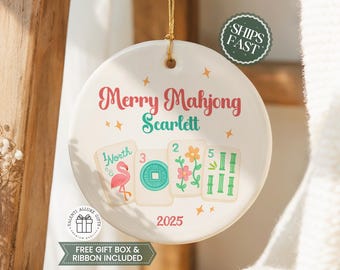 May include: A round white ceramic ornament featuring the text Merry Mahjong and the name Scarlett in red and teal. Below, four mahjong tiles display a pink flamingo, a teal coin, pink flowers, and green bamboo. The year 2025 is printed at the bottom.