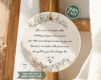 May include: A round white ceramic ornament featuring a gold hanging string and a botanical wreath design with green leaves and small orange berries. The text reads: You are loved for the little girl you once were, for the wonderful woman you've become, and for the precious daughter you'll always be. I LOVE YOU, Mom.