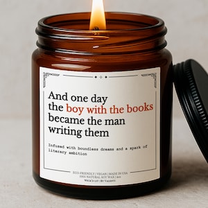 May include: A burning soy wax candle in a brown glass jar with a black lid. The label says "And one day the boy with the books became the man writing them." The candle is 113 grams.