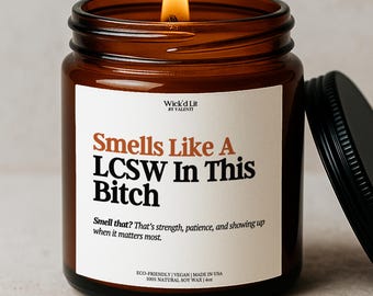 Puede incluir: Un tarro de vela de vidrio ámbar encendido con tapa negra. La vela tiene una etiqueta blanca con el texto "Smells Like A LCSW In This Bitch". La vela está hecha con cera de soja 100% natural y es ecológica y vegana.