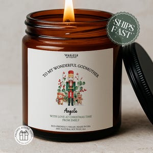 Peut inclure: Une bougie allumée dans un pot en verre brun avec un couvercle noir. L'étiquette indique "TO MY WONDERFUL GODMOTHER" avec une illustration de casse-noisette et le nom "Angela". La bougie est de 113g et fabriquée aux États-Unis.