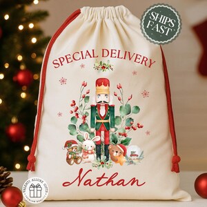 May include: A cream-colored Christmas gift bag with red drawstring and trim. The bag features a nutcracker illustration surrounded by greenery and the words "Special Delivery" and "Nathan" in red. A "Ships Fast" sticker is visible.