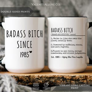 May include: White ceramic mug with a black handle and rim, featuring the text "BADASS BITCH SINCE 1985" on one side and a definition on the other. The mug is double-sided and has a vibrant, long-lasting print.