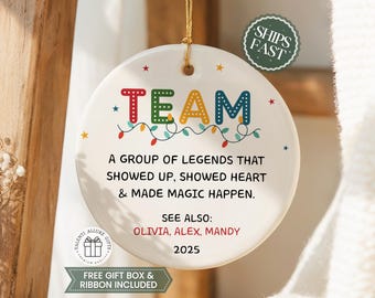 May include: A round white ceramic ornament featuring the word TEAM in colorful letters, a string of Christmas lights, and the text: A GROUP OF LEGENDS THAT SHOWED UP, SHOWED HEART & MADE MAGIC HAPPEN. SEE ALSO: OLIVIA, ALEX, MANDY 2025. It hangs by a gold cord.
