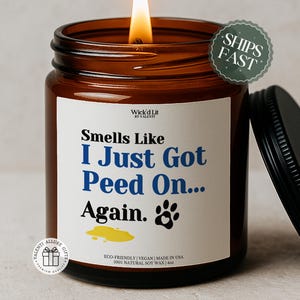 Puede incluir: Una vela encendida en un tarro de vidrio marrón con tapa negra. La vela tiene una etiqueta blanca con el texto "Smells Like I Just Got Peed On... Again." y un gráfico de huella de pata. La vela está hecha con cera de soja 100% natural y pesa 113g.