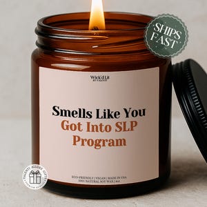Puede incluir: Una vela encendida en un tarro de cristal marrón con tapa negra. La vela tiene una etiqueta blanca con el texto "Smells Like You Got Into SLP Program". La vela está hecha con cera de soja natural y es ecológica.