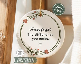 May include: A round white ceramic ornament featuring a green circular vine border with small red flowers. The text reads, "Never forget the difference you make." with a small heart symbol below. It hangs from a thin gold string for display purposes.