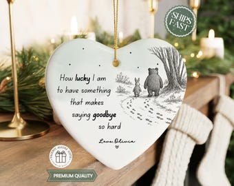 May include: A white heart-shaped ceramic ornament featuring a black ink illustration of a bear and a rabbit walking away on a path. The text reads: How lucky I am to have something that makes saying goodbye so hard. Love, Olivia. It hangs by a gold cord.