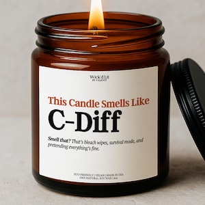 May include: A lit brown glass jar candle with a black lid. The candle has a white label with the text "This Candle Smells Like C-Diff". The label also includes the text "Smell that? That's bleach wipes, survival mode, and pretending everything's fine."