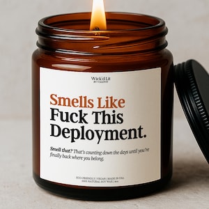 Pode incluir: Uma vela acesa em um frasco de vidro âmbar com tampa preta. A vela tem um rótulo branco com o texto "Smells Like Fuck This Deployment." e um texto menor abaixo. A vela é feita com cera de soja natural.