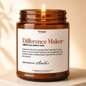 May include: A 4oz amber glass jar candle with a lit wick. The white label features the text Dictionary BY VALENTI, Difference Maker, its definition, and the name Amelia with a small heart icon. The label also states Eco-Friendly, Vegan, Made in USA, and 100% Natural Soy Wax.