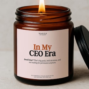 May include: A lit candle in an amber glass jar with a black lid. The label features the text Wick'd Lit by Valenti, In My CEO Era, and Smell that? That's big goals, bold decisions, and not waiting for permission anymore. It states 100% natural soy wax | 4oz.