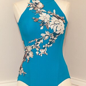 May include: A turquoise bodysuit with a high neckline and halter-style straps. The garment is embellished with a floral design of white sequins and silver beads. The bodysuit is displayed on a mannequin.