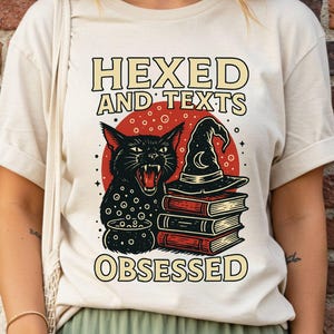 May include: Ivory-colored t-shirt with the words "HEXED AND TEXTS OBSESSED" above a graphic of a black cat, witch hat, and books. The cat is in front of a red circle. The shirt is worn by a person.
