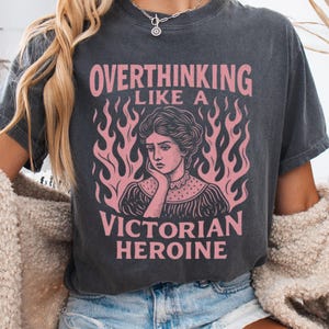 May include: A gray t-shirt with a pink graphic that reads 'Overthinking Like a Victorian Heroine.' The graphic features a Victorian woman with a sad expression surrounded by pink flames. The shirt is worn by a person with blonde hair and is paired with denim shorts.