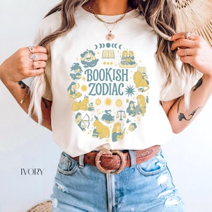 May include: Ivory-colored t-shirt featuring a circular design with the words "Bookish Zodiac" in teal and yellow. The design includes zodiac symbols and illustrations of people reading books. The shirt is paired with blue jeans and a brown belt.