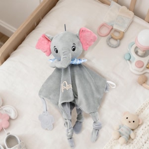 May include: A gray plush elephant-shaped baby security blanket with pink ears, a blue collar, and embroidered initials. The blanket is surrounded by other baby items, including a rattle, a stuffed bunny, a teddy bear, and baby shoes.