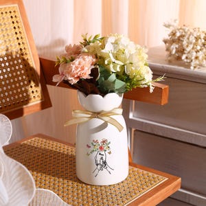 May include: White ceramic vase with a scalloped rim and a gold ribbon. The vase features a hand-drawn design of two hands holding a bouquet of flowers with butterflies. The vase is filled with a bouquet of pink and white flowers.