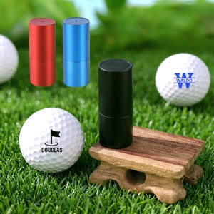 May include: Golf accessories set on green grass. Includes three cylindrical golf ball markers in red, blue, and black. Two golf balls with printed logos and text are also visible. One ball has the name "DOUGLAS" and a flag graphic.