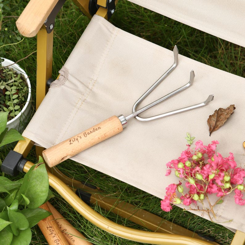 May include: A small garden hand rake with three tines and a wooden handle. The text "Lily's Garden" is engraved on the handle. The rake is resting on a beige chair with pink flowers and green foliage in the background.