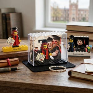 May include: A graduation-themed display featuring Lego figures and a photo. The figures wear graduation caps and gowns, holding diplomas. One figure is in a clear case with a photo of graduates. The display includes a pearl bracelet and a notebook.