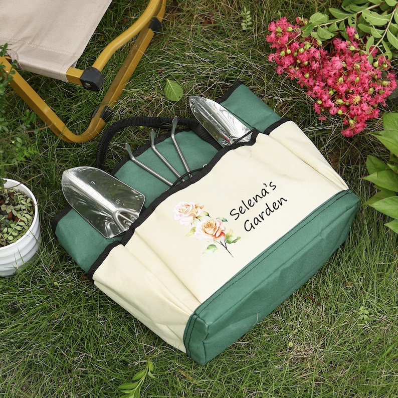 May include: A green and cream-colored gardening tote bag with gardening tools. The bag has a black handle and the text "Selena's Garden" with floral accents. The tools include a trowel, hand rake, and a scoop.