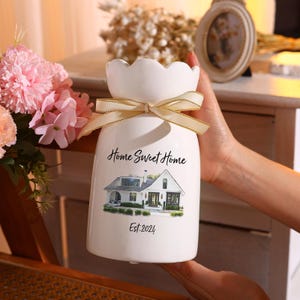 May include: White ceramic vase with a scalloped rim and a gold ribbon bow. The vase features a house illustration with the text "Home Sweet Home" and "Est.2024". The vase is designed to hold flowers.
