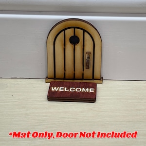 May include: A miniature wooden door with an arched top and a small round window. A rectangular welcome mat with the word "WELCOME" in white lettering is in front of the door. The door is made of light brown wood with dark brown lines.
